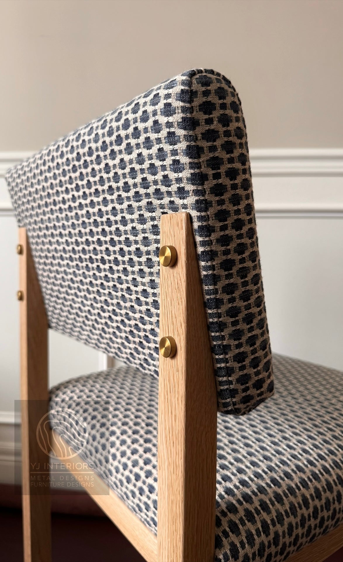A close-up of a wooden dining chair with a patterned fabric upholstery. The fabric features a navy blue and beige geometric dot motif, and the chair has light natural wood legs and gold-colored brass hardware.