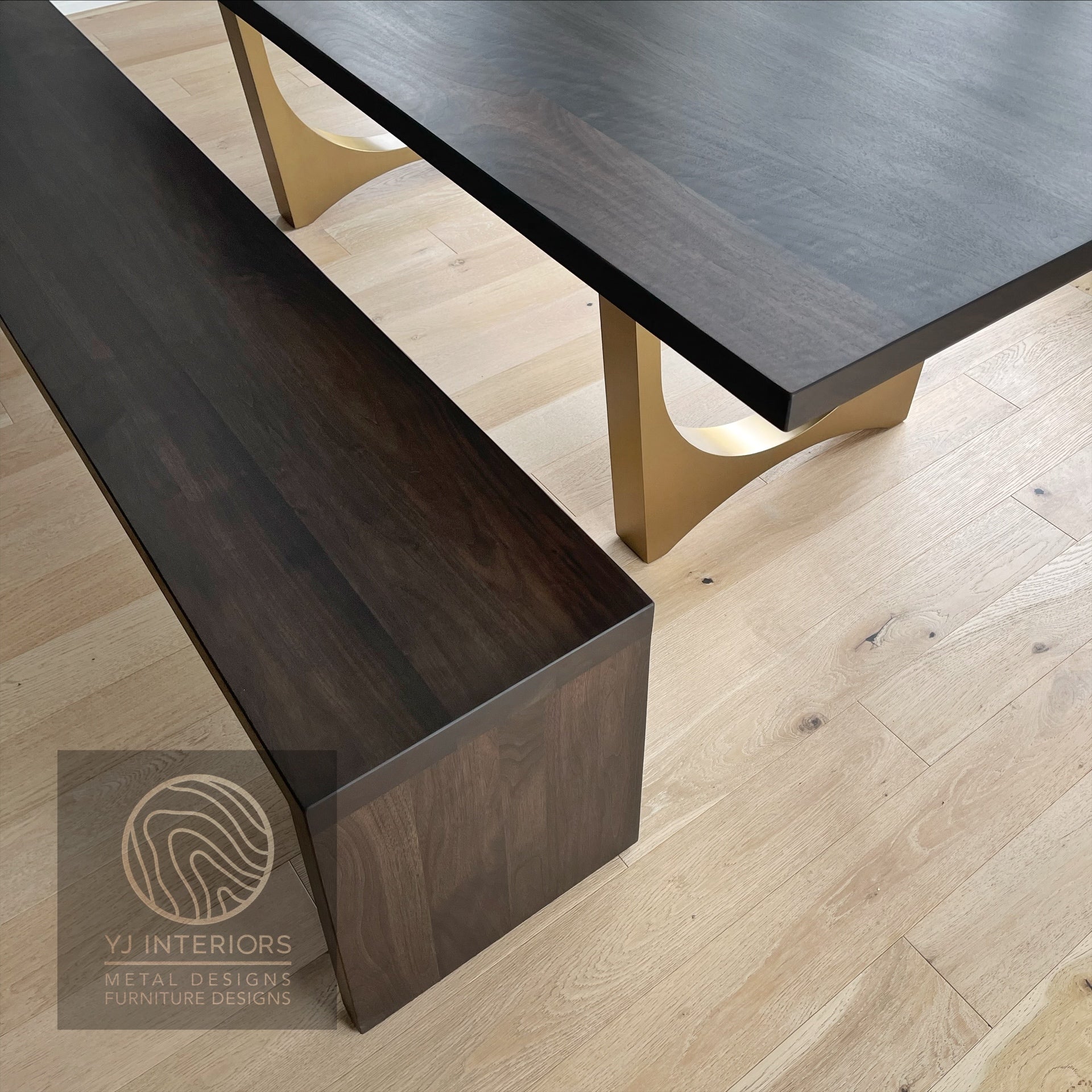 Dark wooden dining table with gold legs on a light wood floor, featuring YJ Interiors logo.