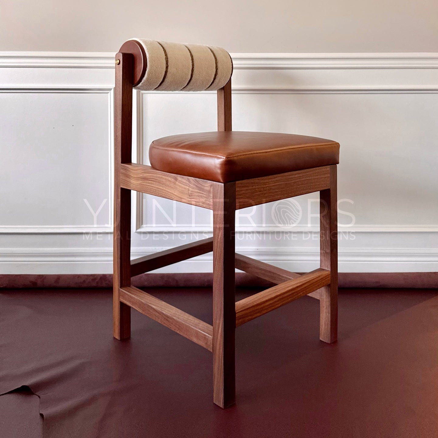 A wooden bar stool with a brown leather seat cushion and a curved backrest upholstered in beige and brown striped fabric, set against a white paneled wall.