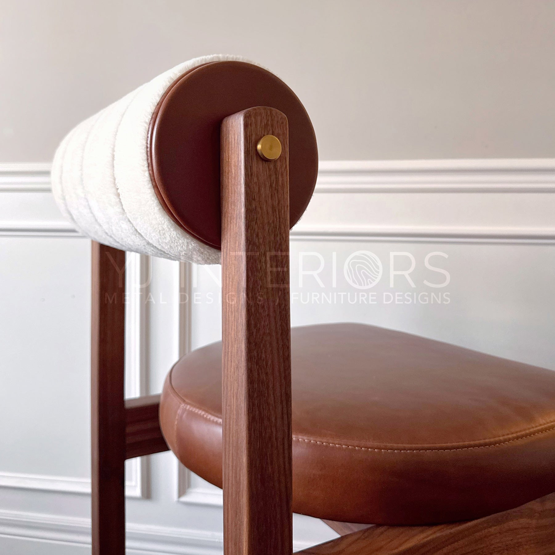 Close-up of a wooden chair with a brown leather seat and backrest, featuring a cylindrical upholstered backrest in cream and brown fabric, set against a light gray wall with white trim.