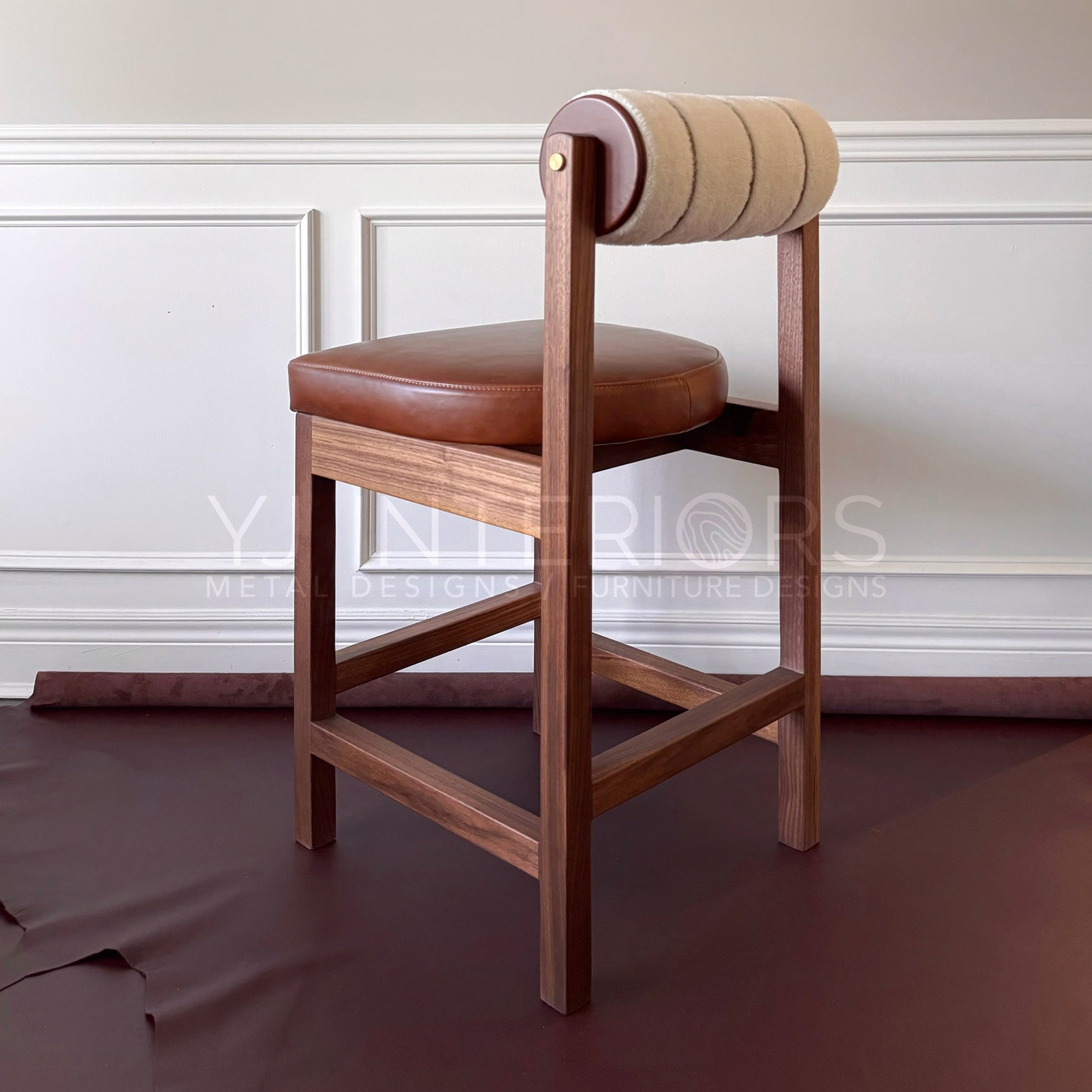 A wooden bar stool with a brown leather seat cushion and a cylindrical upholstered backrest in beige and brown, set against a white paneled wall.