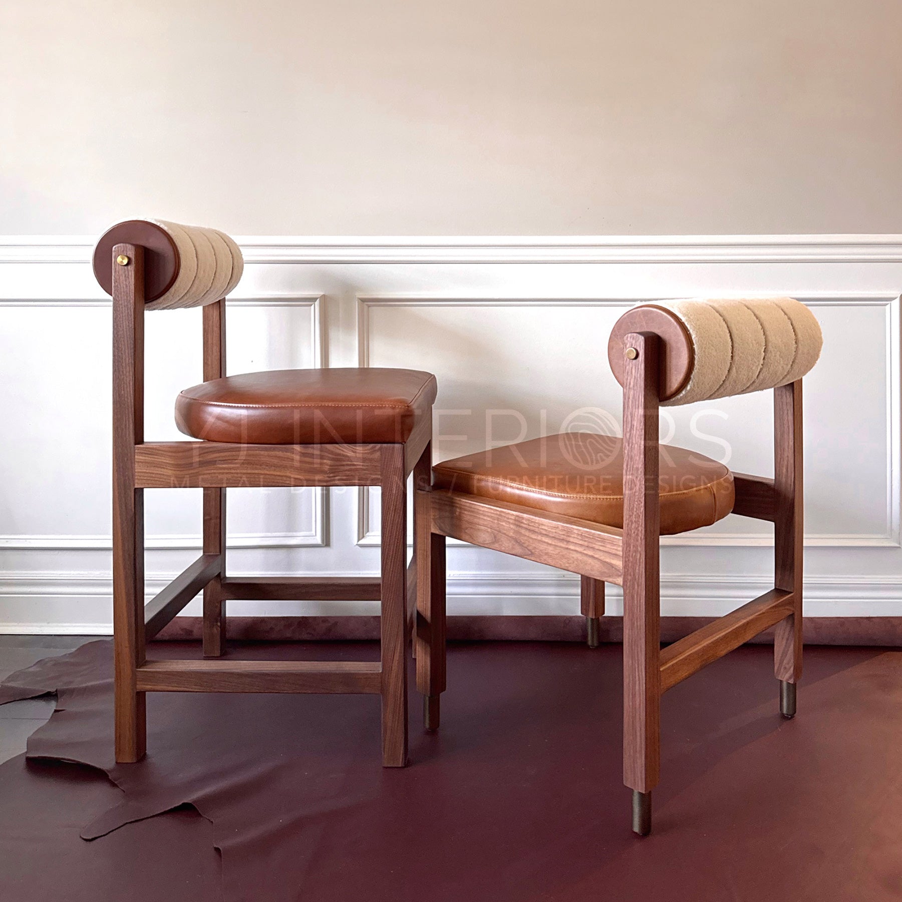 Two wooden dining chairs with brown leather seats and cylindrical beige upholstered backrests, shown in an indoor room with paneled walls and a dark floor.