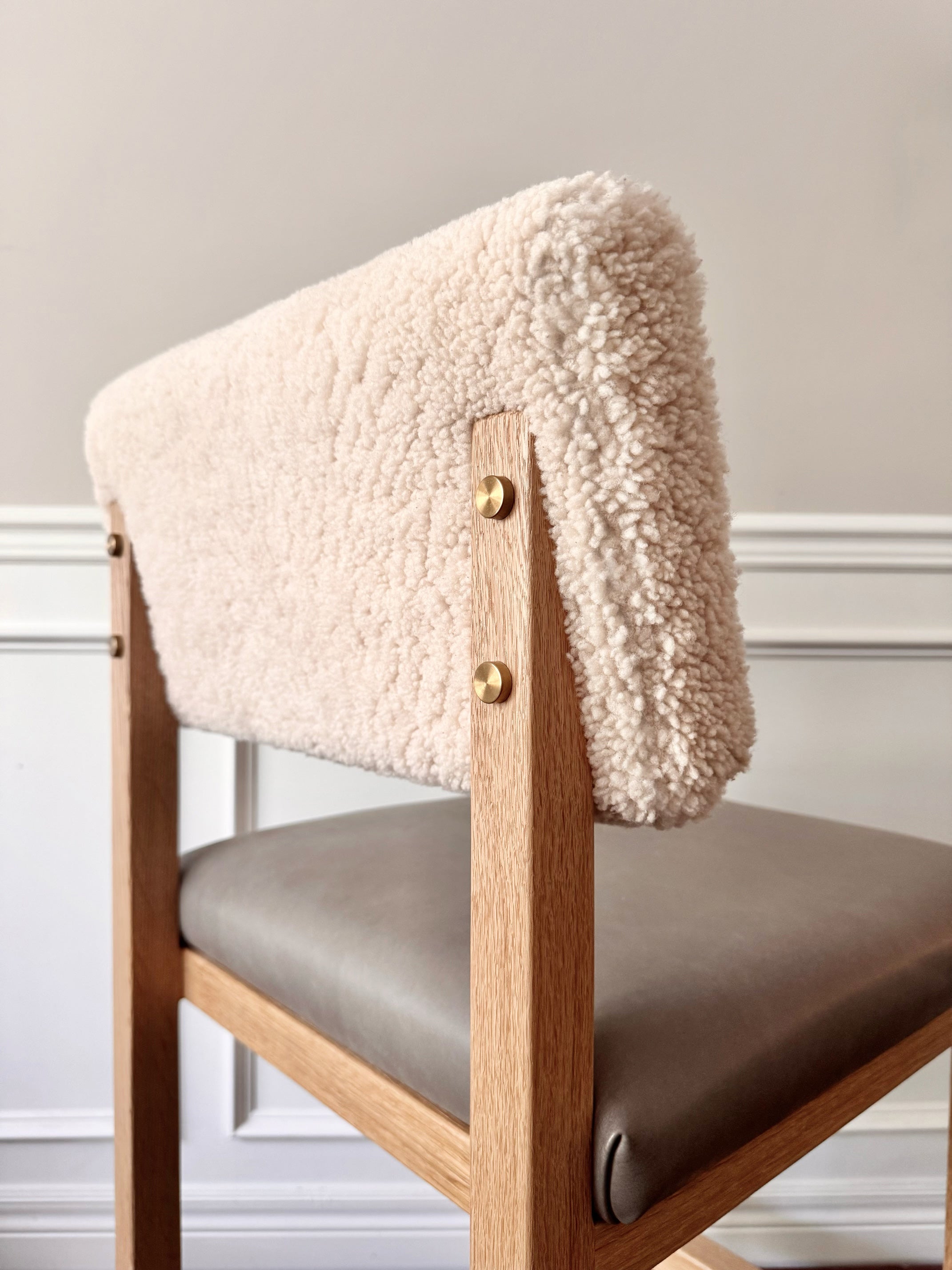 A close-up of a modern wooden chair featuring a thick, textured cream bouclé fabric backrest and a taupe upholstered seat cushion, accented with brass-colored metal hardware.