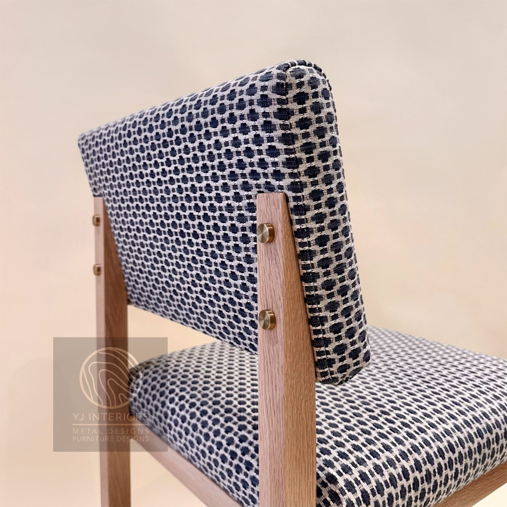 A close-up of a wooden dining chair with a black and white patterned fabric upholstery, featuring brass-colored metal hardware and a light beige background.