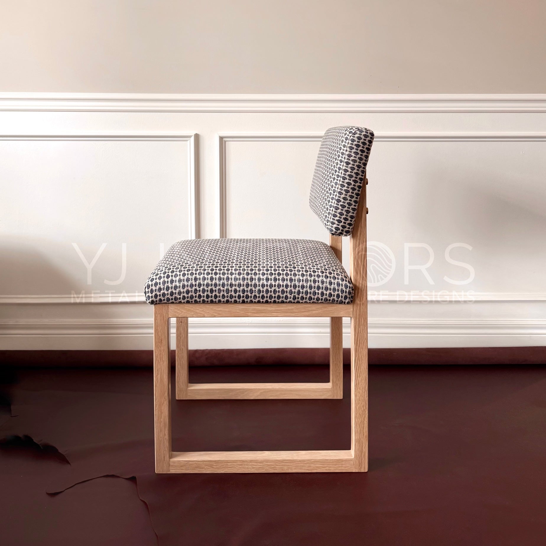A modern wooden dining chair with a light oak frame and a black-and-white geometric patterned cushion, set against a cream paneled wall and dark reddish-brown floor. 